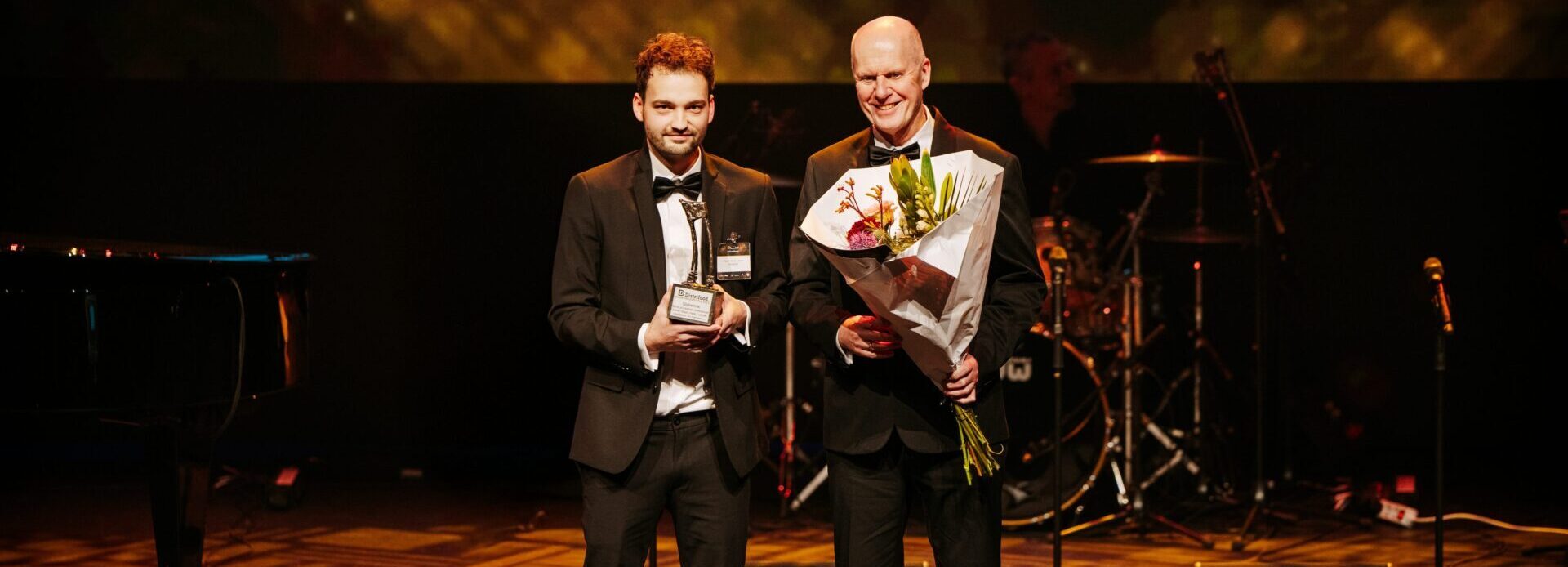 Twee mannen in gala-kleding staan op een podium tijdens een prijsuitreiking. De jongere man links houdt een trofee vast, terwijl de oudere man rechts een bos bloemen vasthoudt. Ze lachen en kijken naar de camera. Op de achtergrond is een bandopstelling met een drumstel en microfoons zichtbaar. Het podium is verlicht met warme, gouden verlichting.